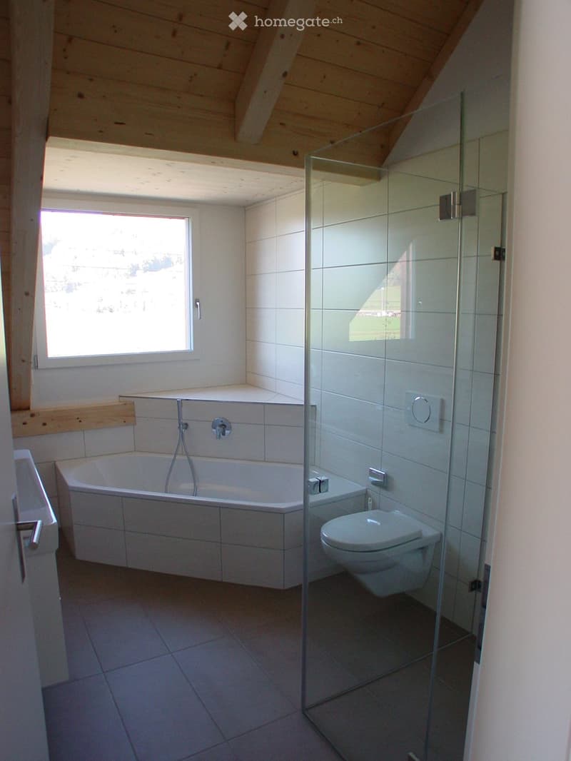 Modern bathroom with a bathtub, toilet, and glass shower enclosure. The room features white tiled walls and a wooden slanted ceiling.