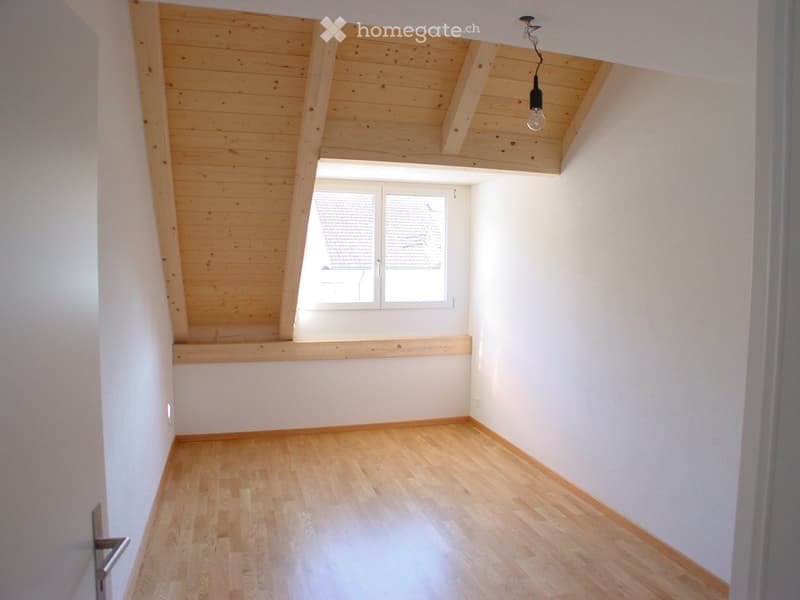 Empty attic room with wooden ceiling, wooden floor, and large window with blinds.