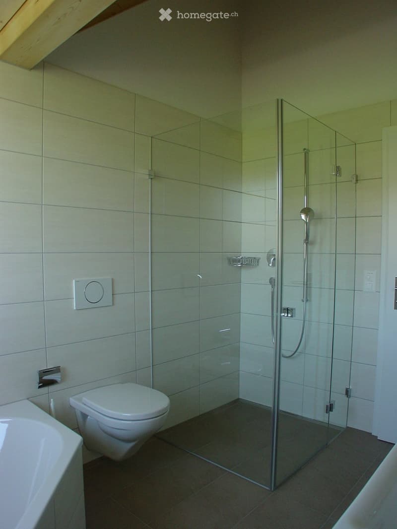 Modern bathroom with a white toilet, bathtub, and glass-enclosed shower with a modern shower handle. The walls are tiled in light gray, and the floor is dark gray tile.