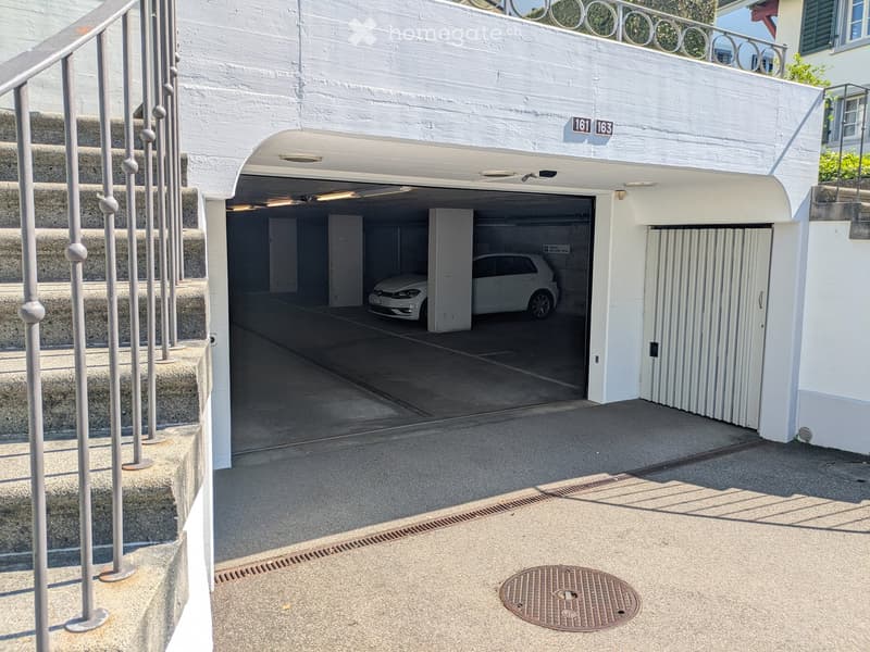 White building with a covered parking area, featuring a white car parked inside and a staircase leading up to a higher level.