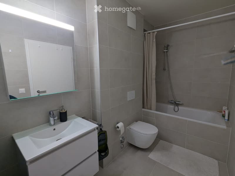 Modern bathroom with white tiles, a white sink, a mirror, a toilet, and a bathtub with a shower curtain. The sink has a faucet and a soap dispenser. There is a towel on the towel rack and a power outlet on the wall.