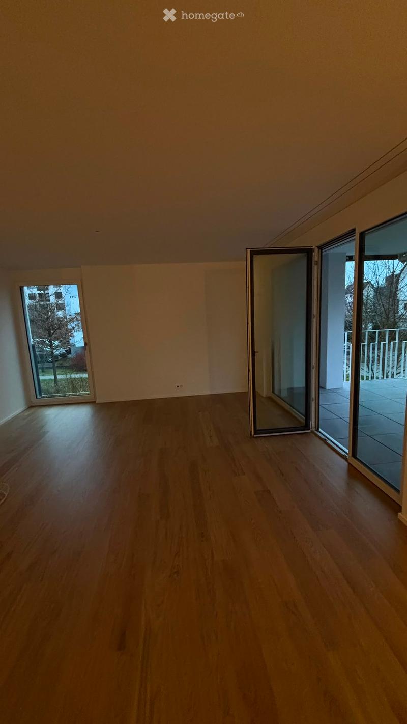 Empty room with wooden flooring, large windows, and sliding glass doors leading to a balcony.
