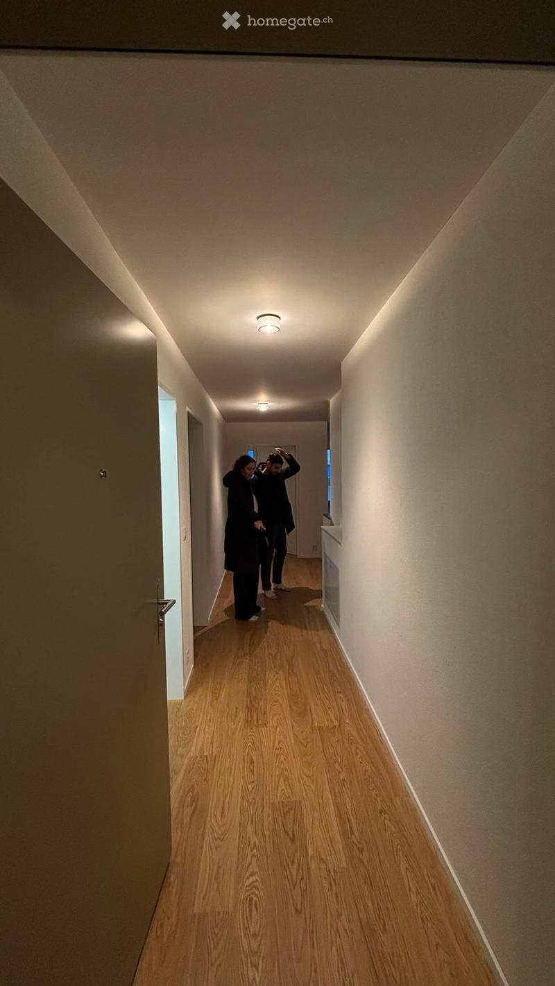 A narrow hallway with wooden flooring, beige walls, and recessed ceiling lighting. Two individuals are walking down the hallway.