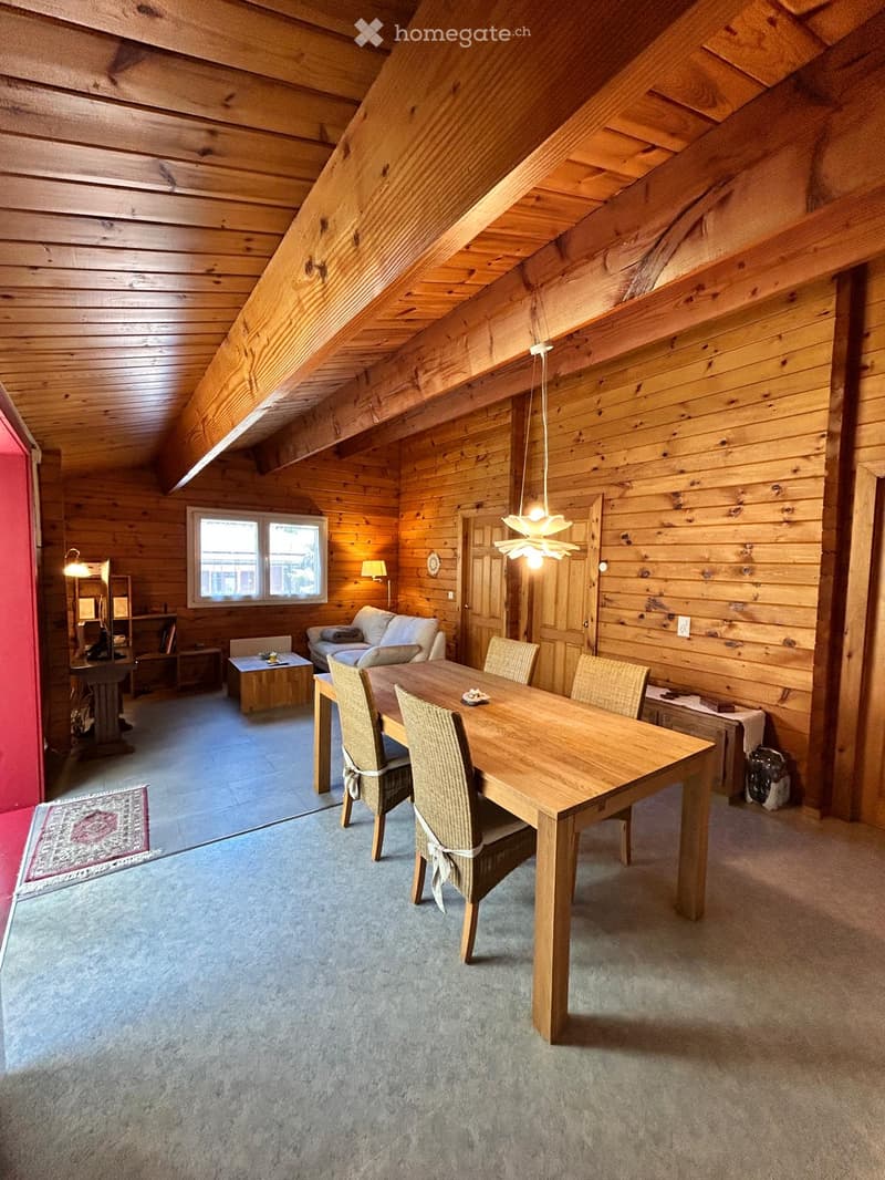 Cozy, wooden-paneled room with a dining table, chairs, sofa, and a lamp. The room features exposed wooden beams on the ceiling and a large window letting in natural light.