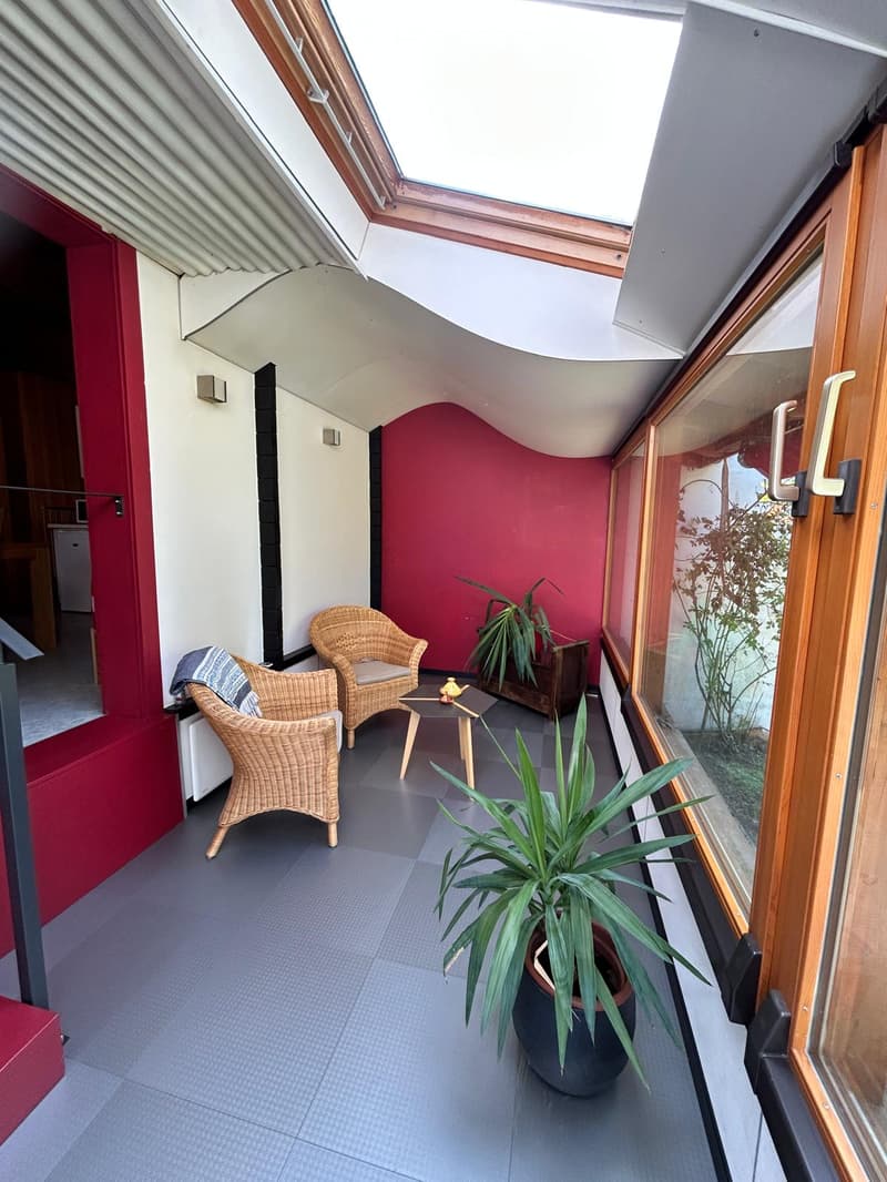 Balcony with grey tiled floor, red accent wall, two wicker chairs, small table, and potted plants.