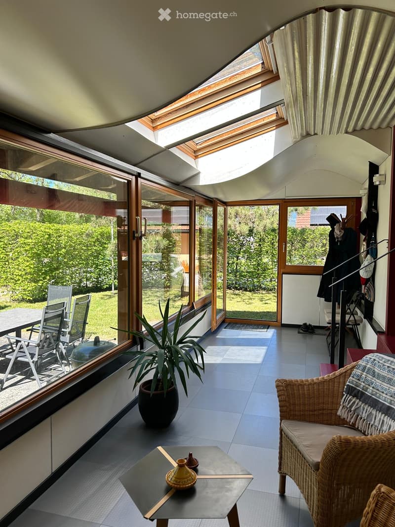 Bright, modern living space with large windows and a skylight, featuring a wicker chair, a small table with a potted plant, and a coffee table with decorative items.