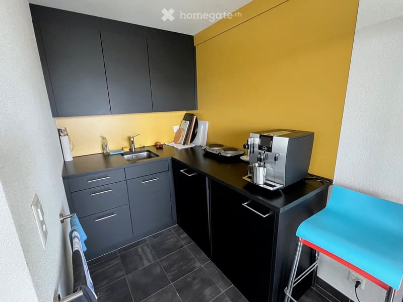 Modern kitchen with dark grey cabinets, black countertops, a sink, a coffee machine, and a stool. The walls are painted in a bright yellow color, and the floor is tiled in dark grey.