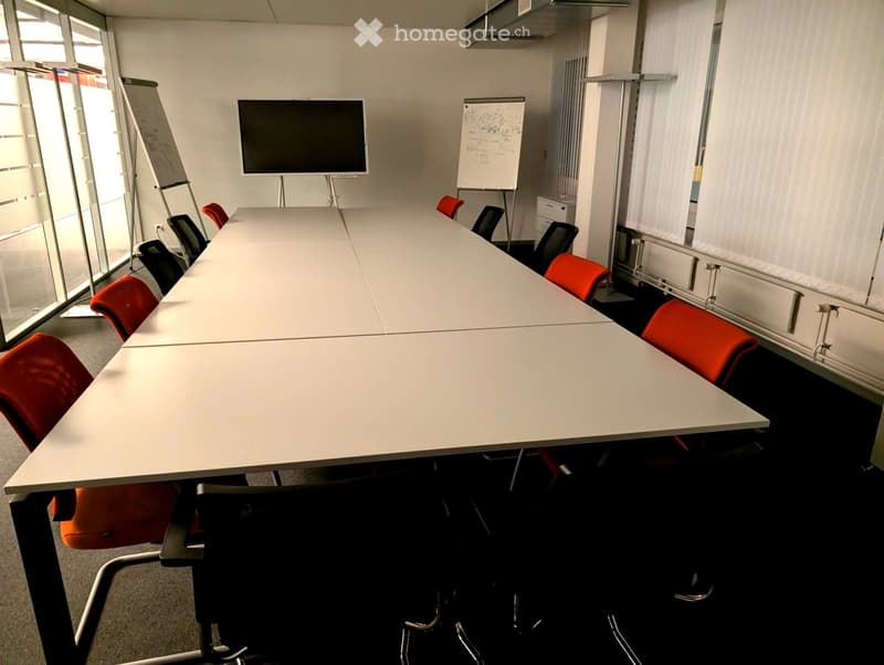 Conference room with a long white table, red and black chairs, a large screen, and a whiteboard.