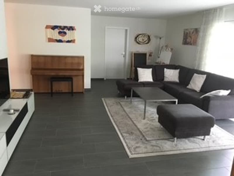 Modern living room with dark tiled flooring, grey sectional sofa, coffee table, ottoman, piano, wall-mounted TV, side table, and wall art.