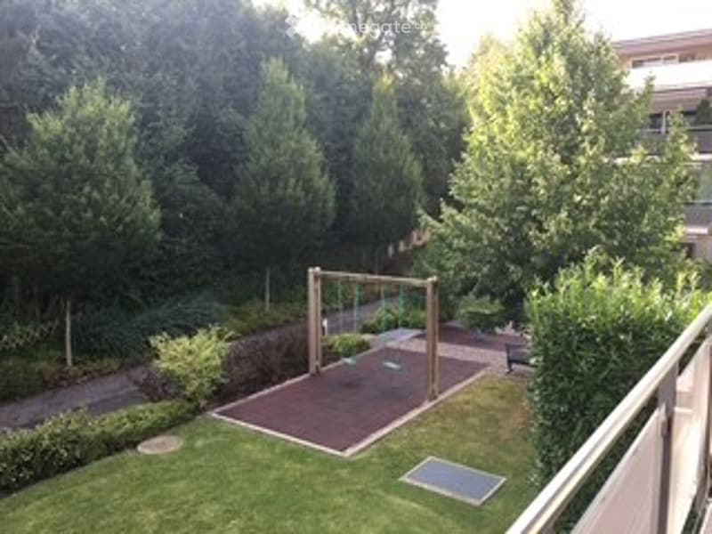A small, fenced backyard with a playground structure, grassy area, and a paved patio. A balcony overlooks the yard.