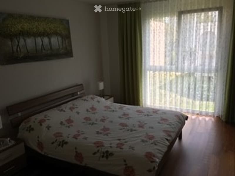 Cozy bedroom with wooden flooring, large window with sheer curtains, a bed with floral bedding, a nightstand with a lamp, and a painting on the wall.