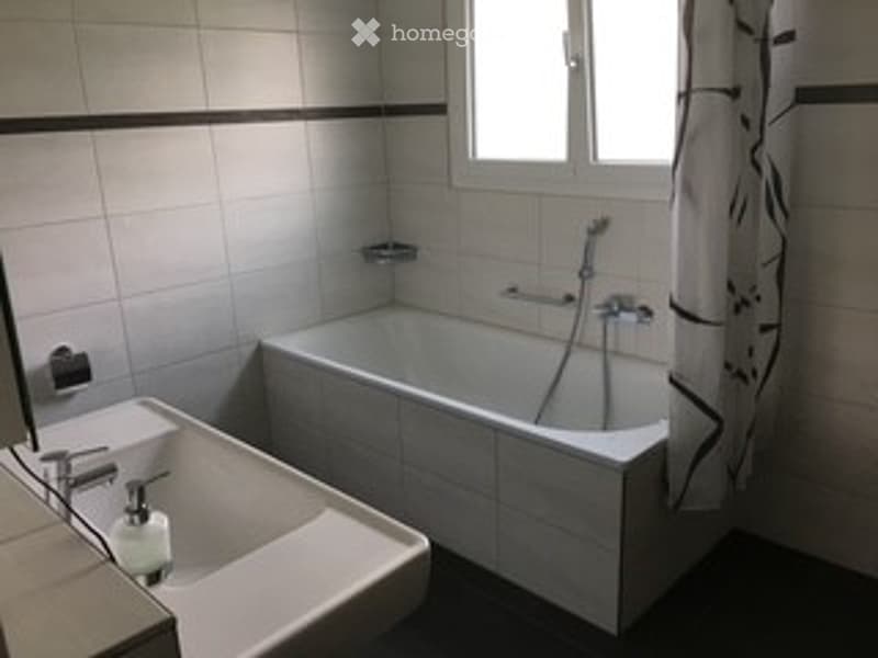 A clean, modern bathroom with a bathtub, toilet, and a window with a curtain. The walls are tiled in white with black trim, and the floor is dark.