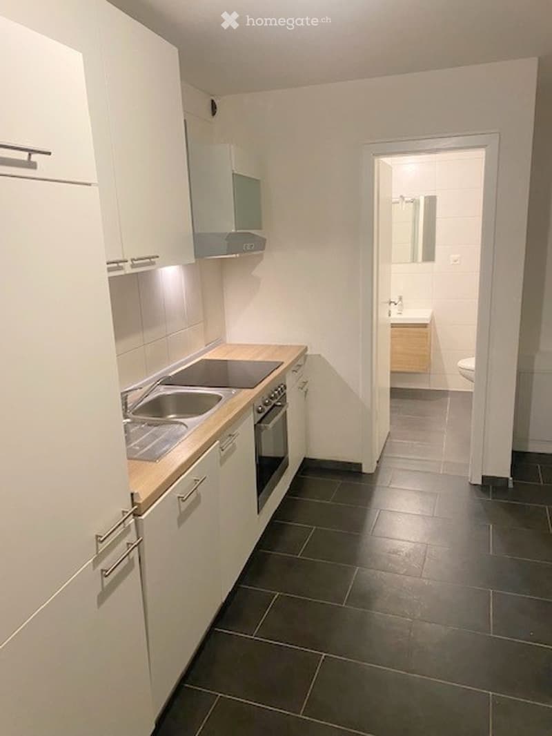Modern kitchen with white cabinets, stainless steel sink, oven, and a small bathroom visible through an open doorway.