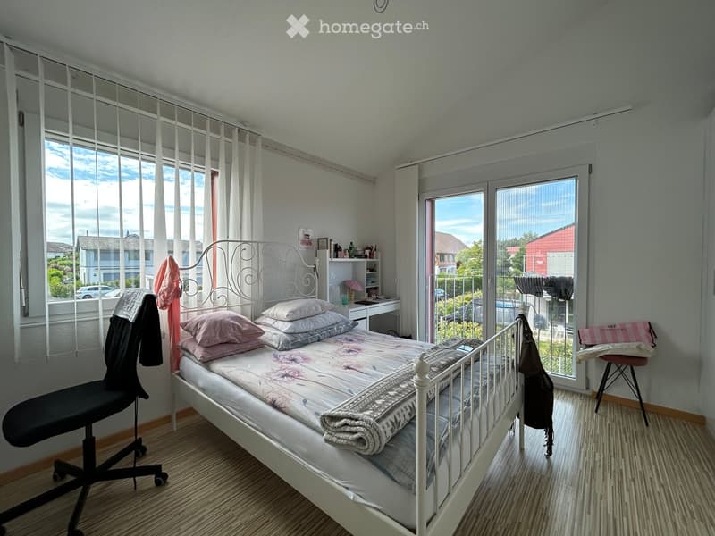 Bright bedroom with a double bed, pillows, and a patterned bedspread. A chair is positioned near the bed, and a small desk with a mirror is visible. The room has large windows with white curtains and a sliding glass door leading to a balcony.