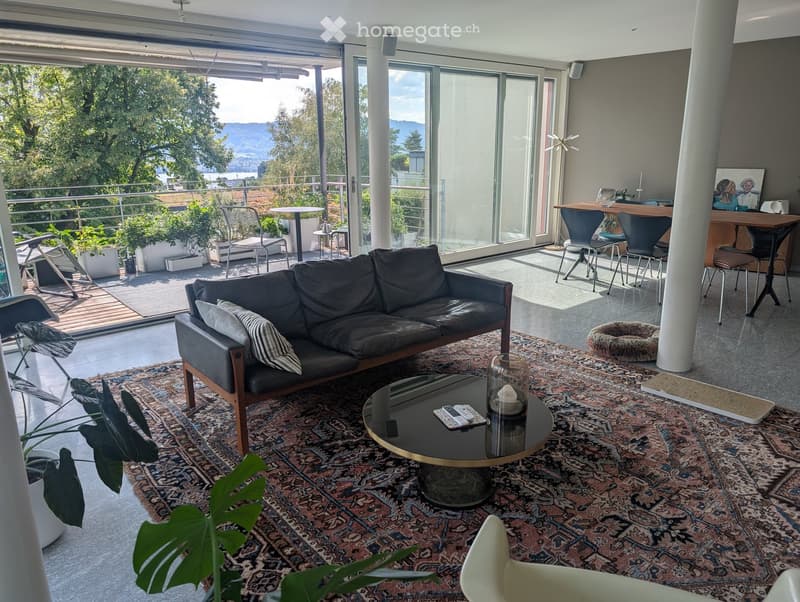 Modern living room with large windows offering a scenic view, featuring a dark leather sofa, coffee table, chairs, and a small table with stools in the adjacent area.