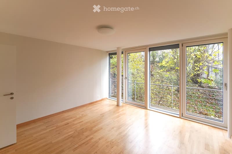 Empty room with light wooden flooring, white walls, and large sliding glass doors leading to a balcony with a view of greenery.