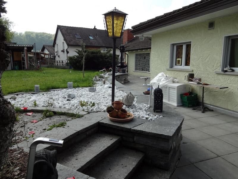 Garden area with a stone pathway, decorative white pebbles, a small table with ceramic pots, a lantern, and a small white cabinet. The background shows a house with a tiled roof and a grassy lawn.