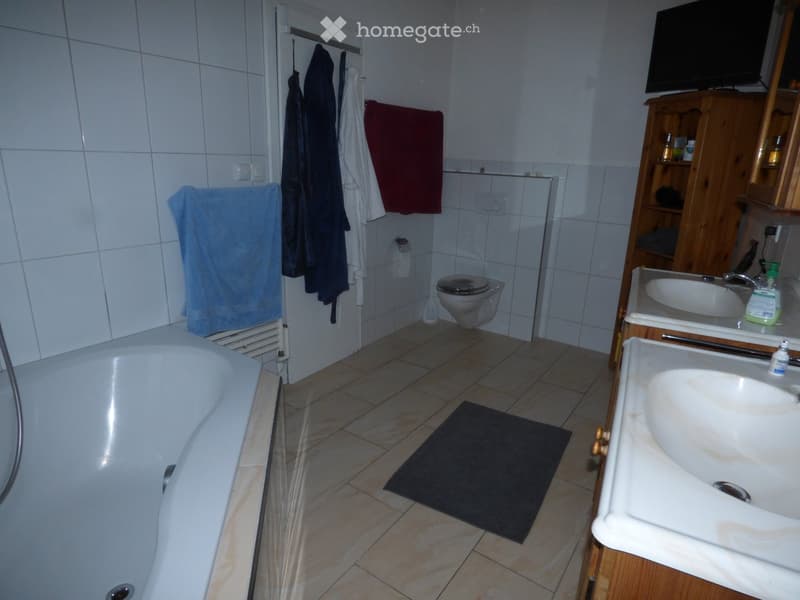 Bathroom with a bathtub, toilet, sink, mirror, and wooden cabinet. Towels are hanging on the wall, and a black bath mat is on the floor.