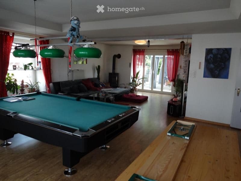 A spacious living room with a pool table, a sofa, a coffee table, a fireplace, and potted plants. The room features large windows with red curtains and green hanging lamps.