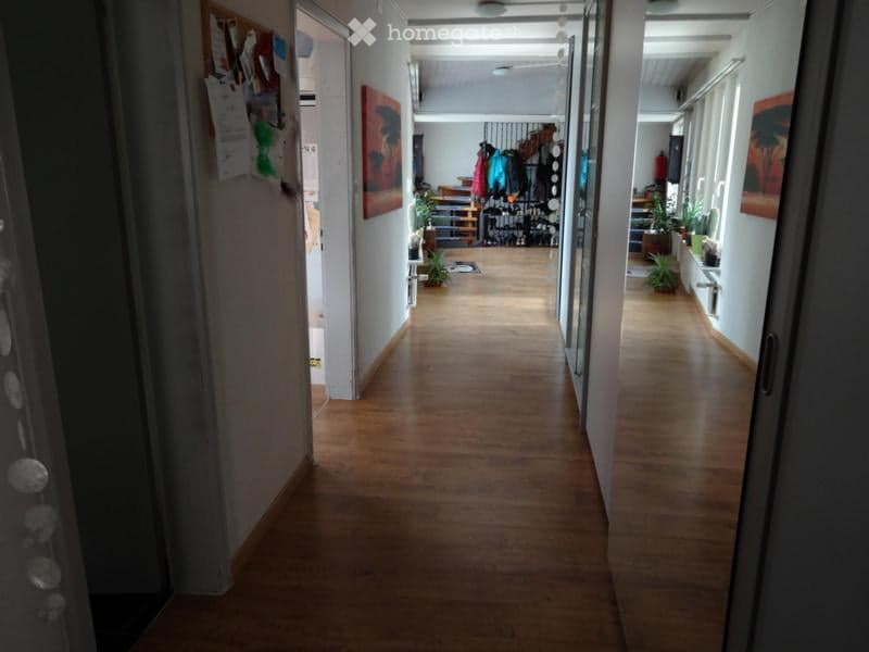 A long, well-lit hallway with wooden flooring, white walls, and several doors leading to different rooms. There are small plants and artwork visible along the walls.