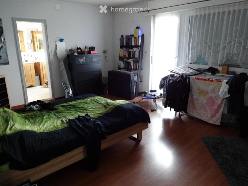 Bedroom with wooden flooring, a bed with green and black bedding, a wardrobe, a bookshelf, a mirror, a chair, and a clothing rack with hanging clothes.