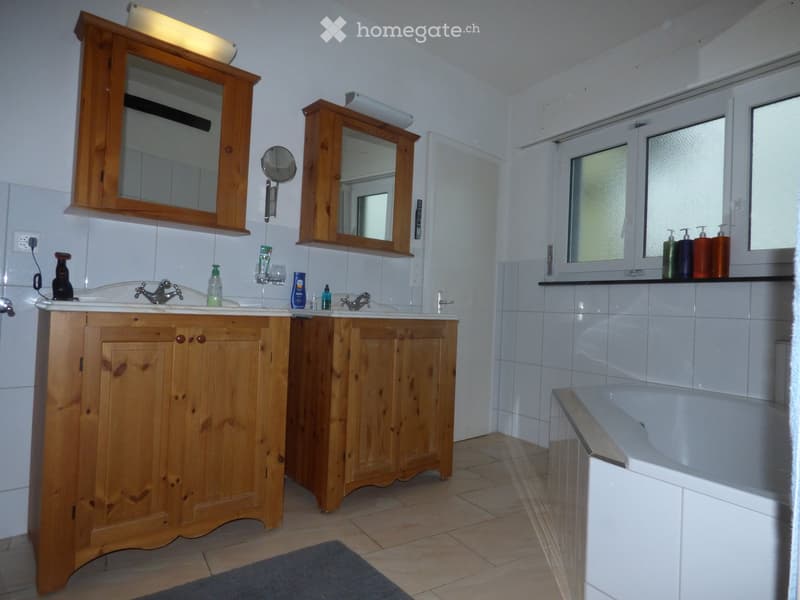 Bathroom with double sink, wooden cabinet, mirrors, bathtub, and toiletries on the windowsill.