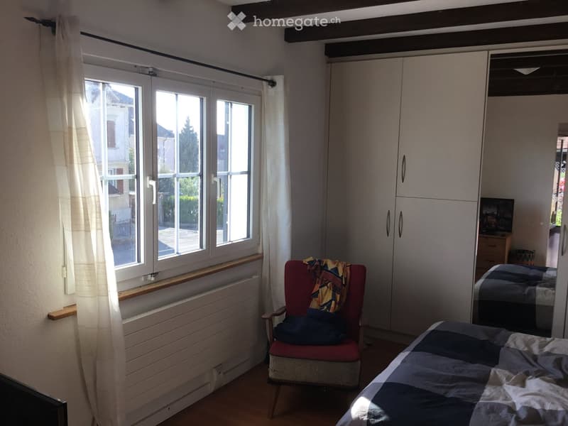 Bright bedroom with wooden flooring, large window with white curtains, a red armchair with a blanket, a bed with a patterned quilt, and a white wardrobe with mirrored doors.