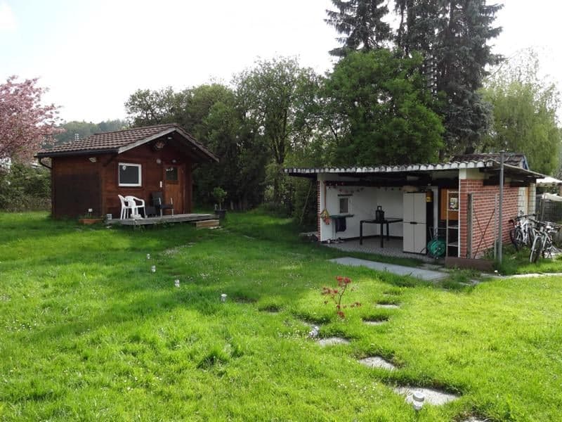 A grassy outdoor area with a wooden cabin and a small utility shed. The cabin has a porch with chairs and a table, while the shed contains a refrigerator, sink, and other outdoor equipment.