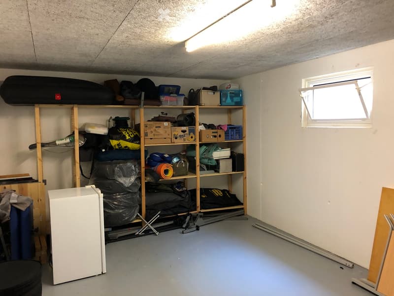 Storage room with wooden shelves filled with various items including bags, boxes, and tools. A small refrigerator is visible on the left side.