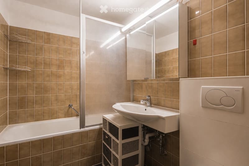 Modern bathroom with beige tiled walls, a white bathtub, a sink with a chrome faucet, a large mirror, and a storage unit beneath the sink.