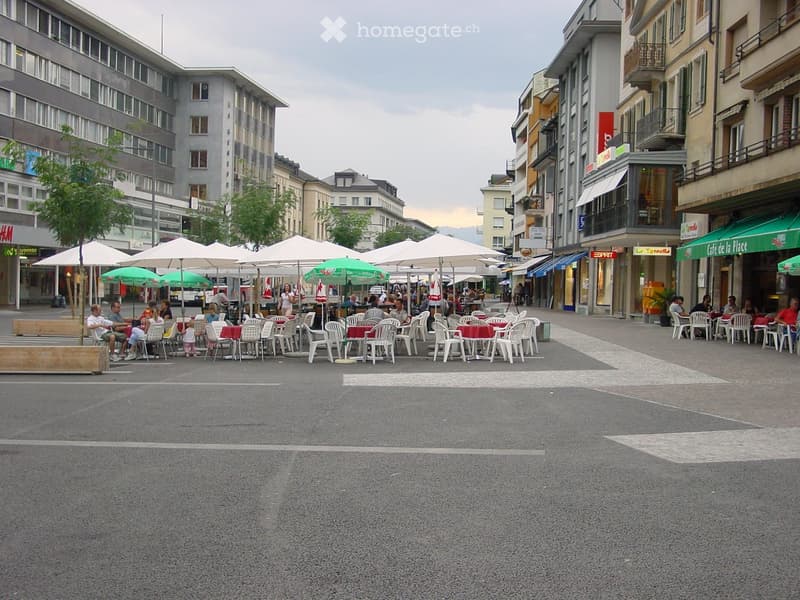 An outdoor seating area with tables and chairs under umbrellas, surrounded by buildings and shops.