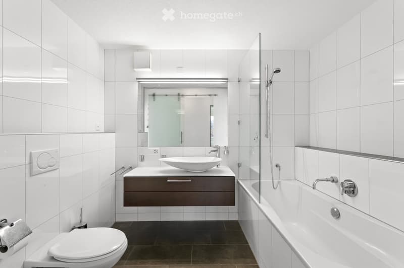 Modern bathroom with white tiles, a white sink with a dark wooden cabinet, a bathtub with a glass shower enclosure, and a toilet. The space features sleek fixtures and a minimalist design.