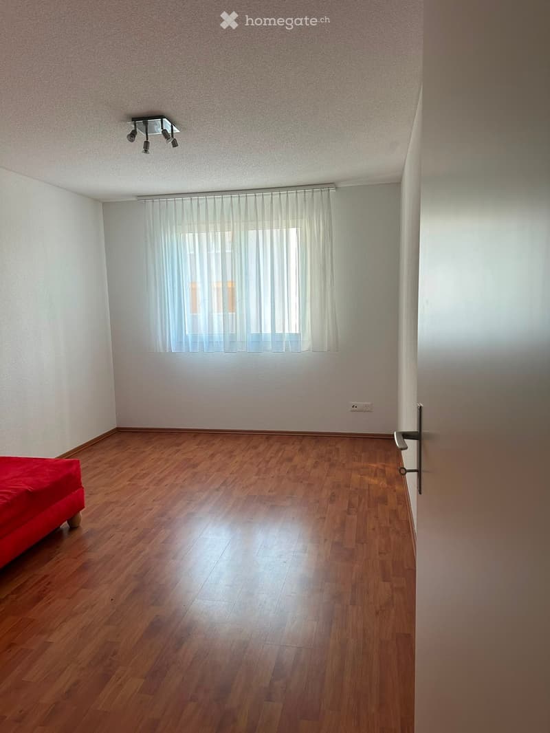 Empty bedroom with wooden flooring, white walls, and a red bed visible on the left side.