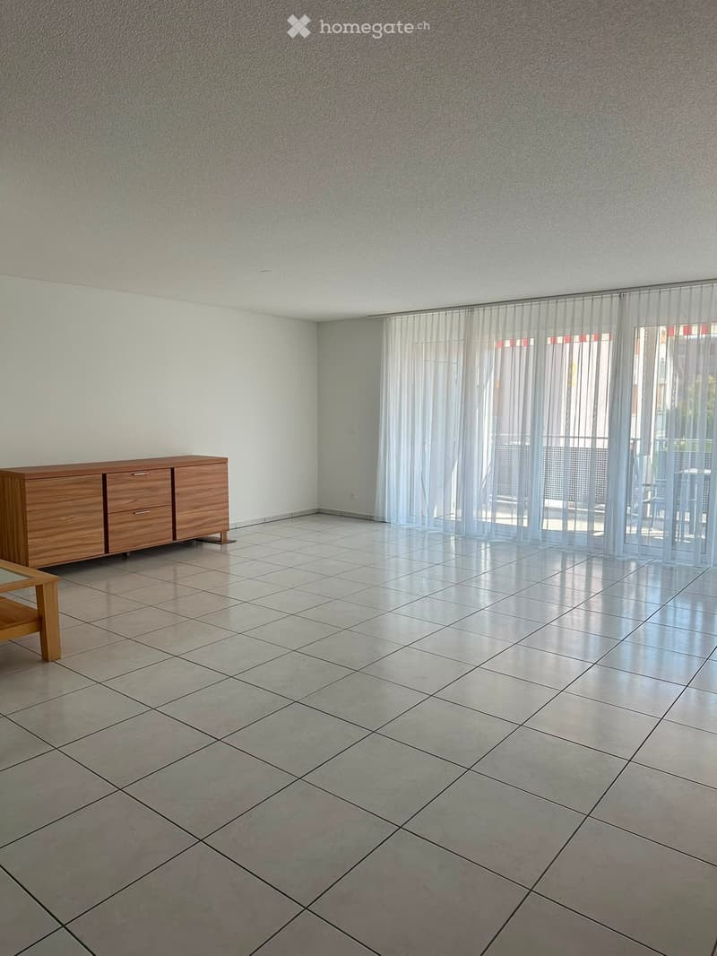 A furnished room with a tiled floor, a wooden cabinet, and a small wooden table near the window with sheer curtains.