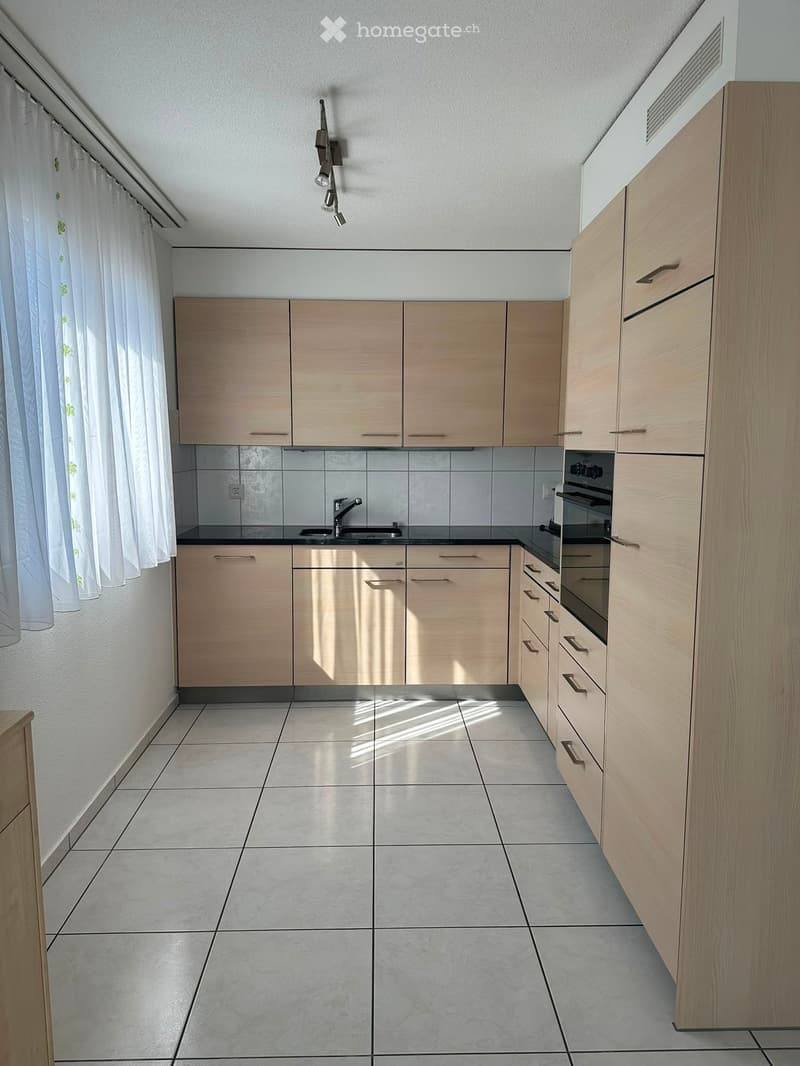 Modern kitchen with light wooden cabinets, a sink, and an oven. The space features tiled flooring and white walls with a window covered by a white curtain.