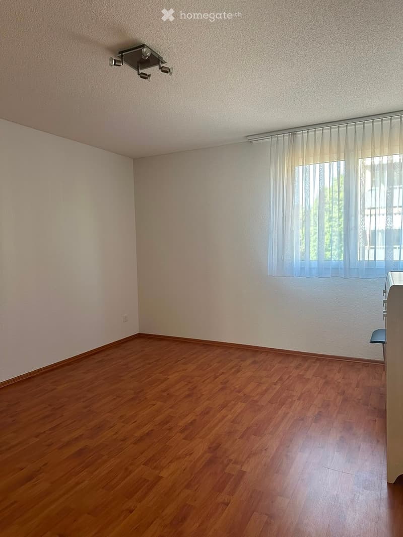 Empty room with wooden flooring, white walls, and a window with sheer curtains. A small portion of a desk is visible on the right side.