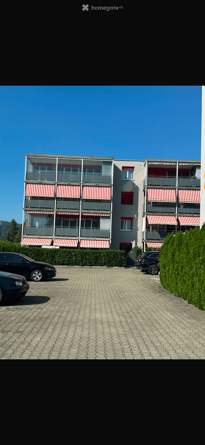 Modern multi-story building with balconies, striped awnings, and a paved parking area.