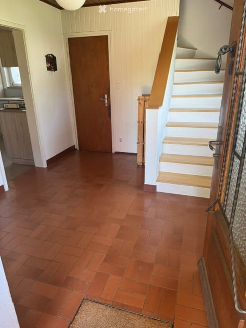 Spacious hallway with polished wooden flooring, white walls, and a wooden staircase leading upwards. Features a closed wooden door and a small decorative item on the wall.