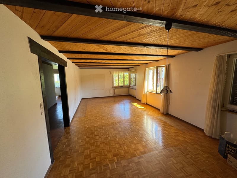 Empty room with wooden flooring, exposed wooden beams on the ceiling, large windows with curtains, and a radiator under one window.