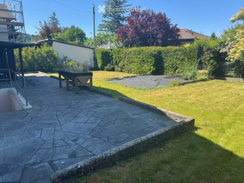 Well-maintained garden with a stone patio, wooden bench, neatly trimmed hedges, and a small grassy area.