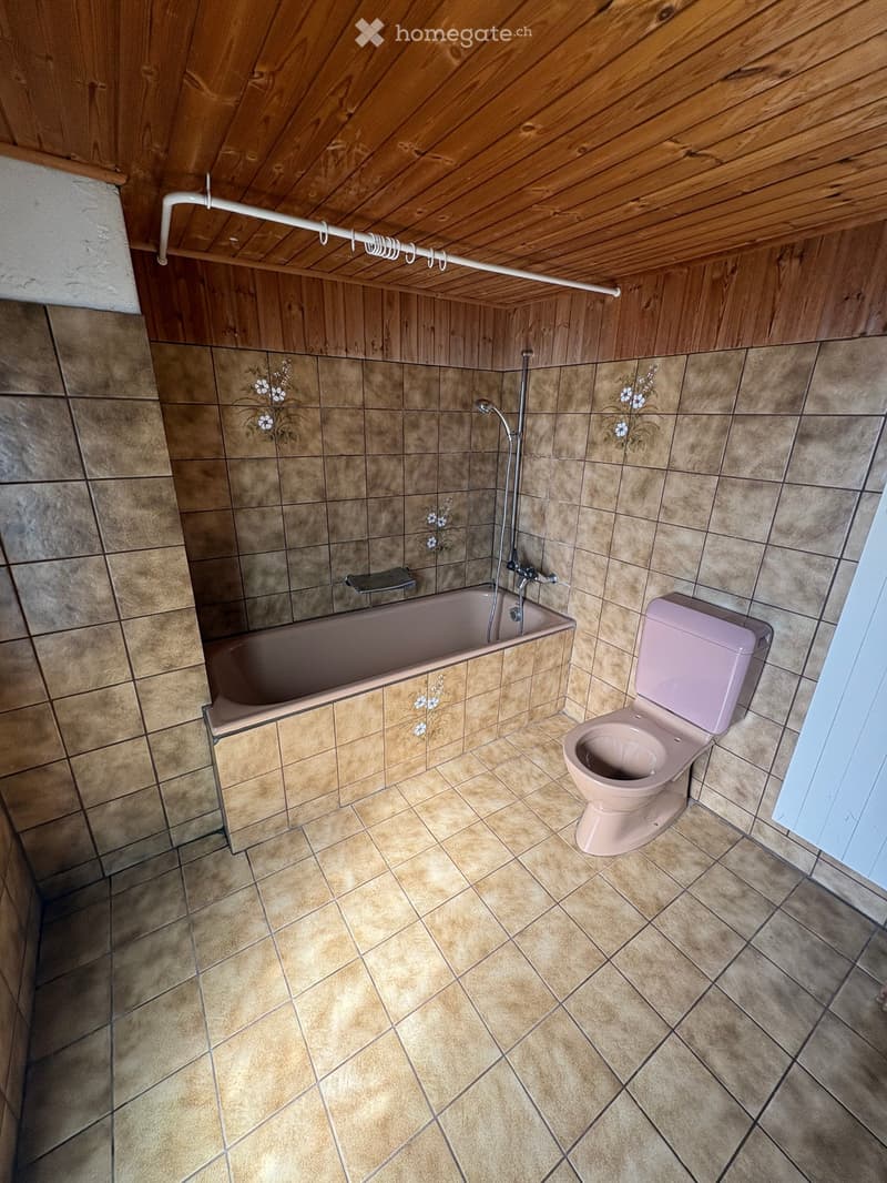 Bathroom with beige tiled walls and floor, featuring a pink bathtub and toilet, wooden ceiling, and floral tile designs.