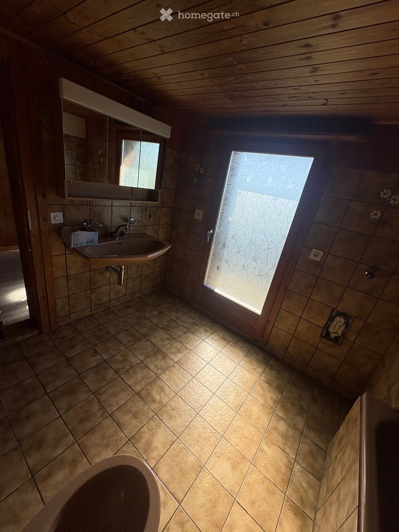Bathroom with tiled walls and floor, a sink with a mirror above it, and a bathtub. The room features wooden beams on the ceiling and a frosted window letting in natural light.