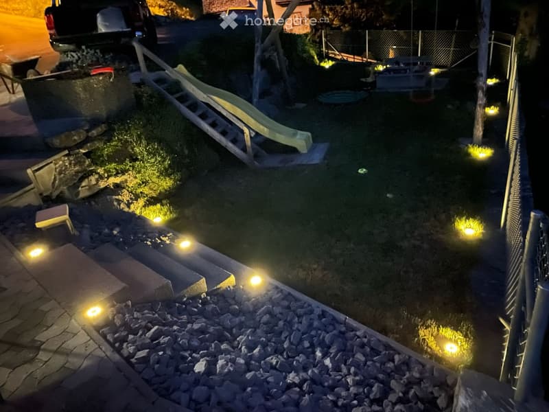 Nighttime view of a small garden area with a pool, surrounded by small lights, a lounge chair, and a fence.