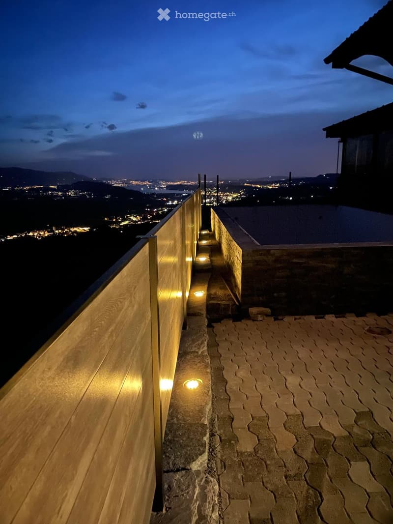 Wooden railing with integrated lighting, stone-paved terrace, cityscape view in the background.