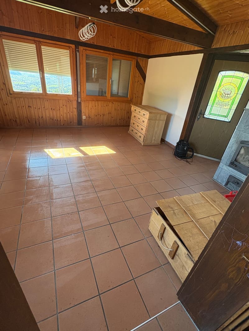 Empty room with tiled floor, wooden walls, and large windows. Features include a wooden chest of drawers, a small wooden bench, and a stained-glass window.