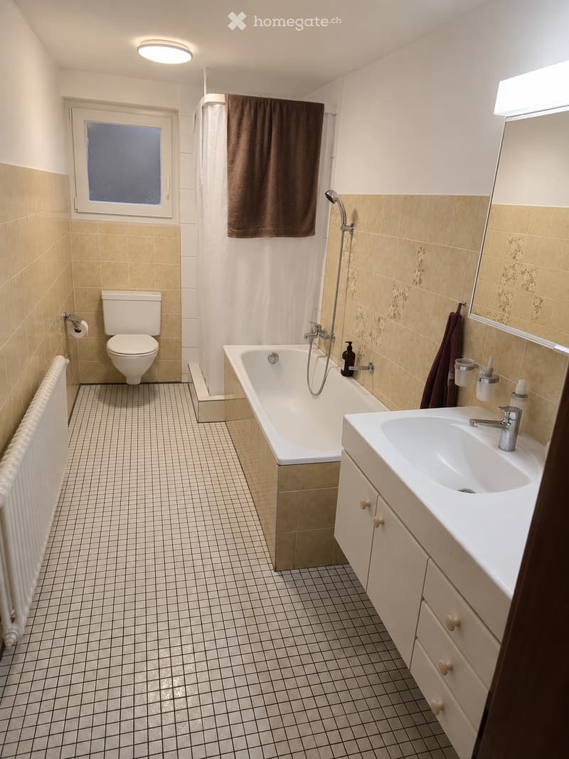 Bathroom with beige tiled walls, white bathtub, toilet, sink, and mirror. Features a shower hose, towel, and radiator.