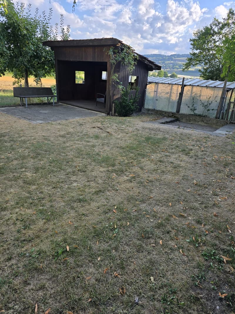 A small wooden outdoor structure with a bench and a chair, surrounded by grass and trees, with a view of a field and hills in the background.