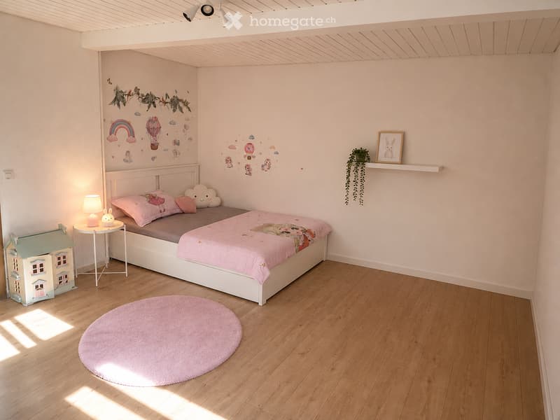Cozy children's bedroom with a white bed, pink bedding, a round pink rug, a small nightstand with a lamp, a toy house, and wall decals featuring clouds and rainbows.