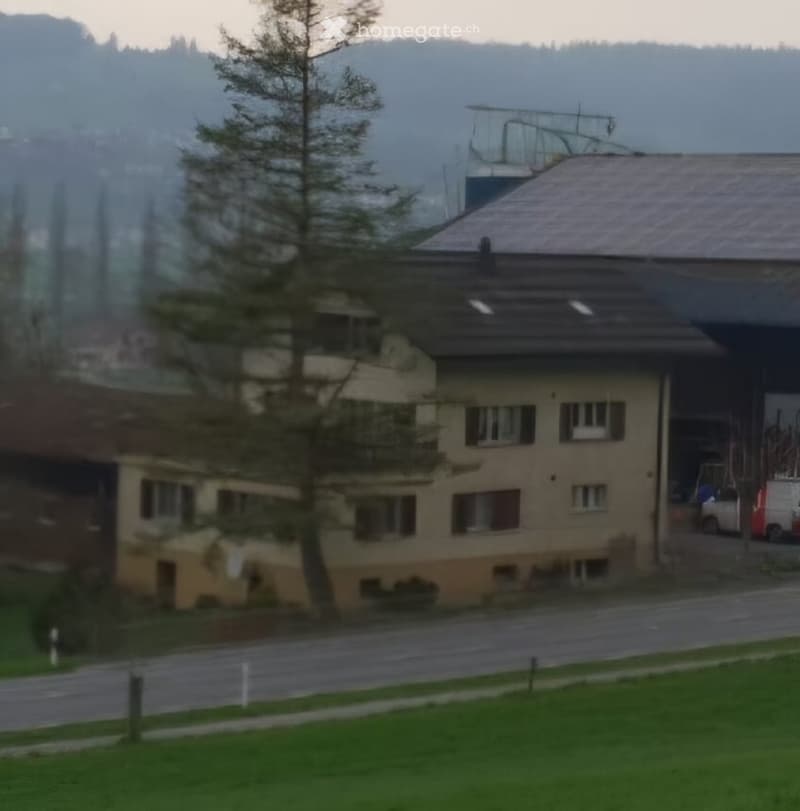 A multi-story residential building with a sloped roof, multiple windows, and a visible garage entrance. The building is surrounded by a grassy area and trees, with a road in front.