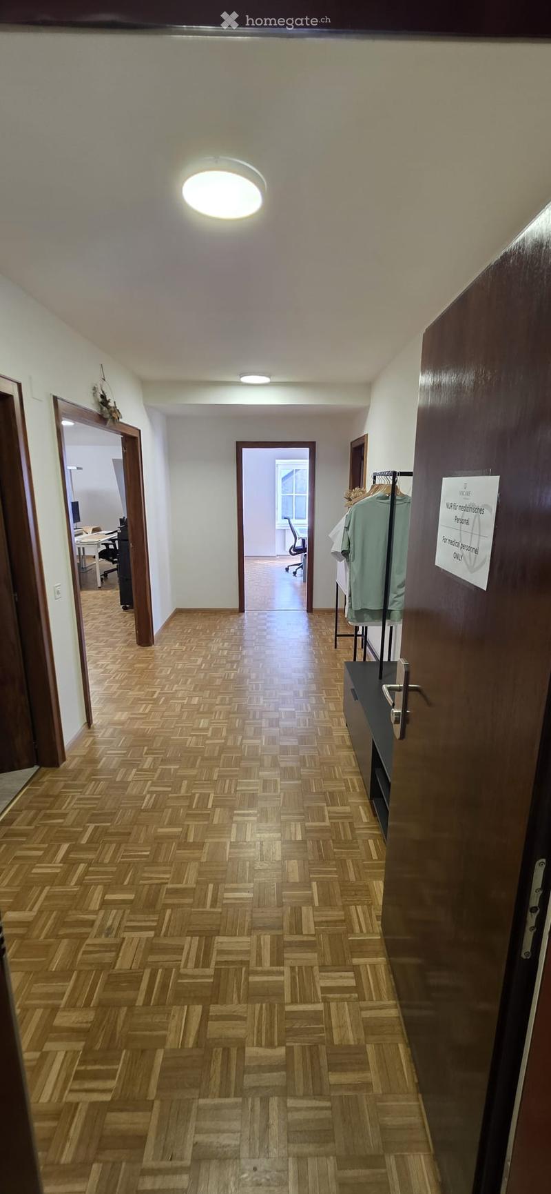 A long, narrow hallway with wooden herringbone flooring, white walls, and recessed lighting. Doors lead to adjacent rooms, including an office area visible in the background.